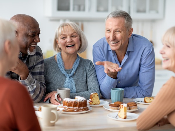 Multiracial group of happy senior people sitting around table drinking tea with cake and having conversation, smiling and laughing, having home party or enjoying time at nursing home