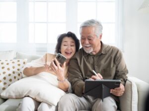 Asian senior couple in living room at home.Wife browsing online on smartphone showing something to her husband while husband is also using a tablet.